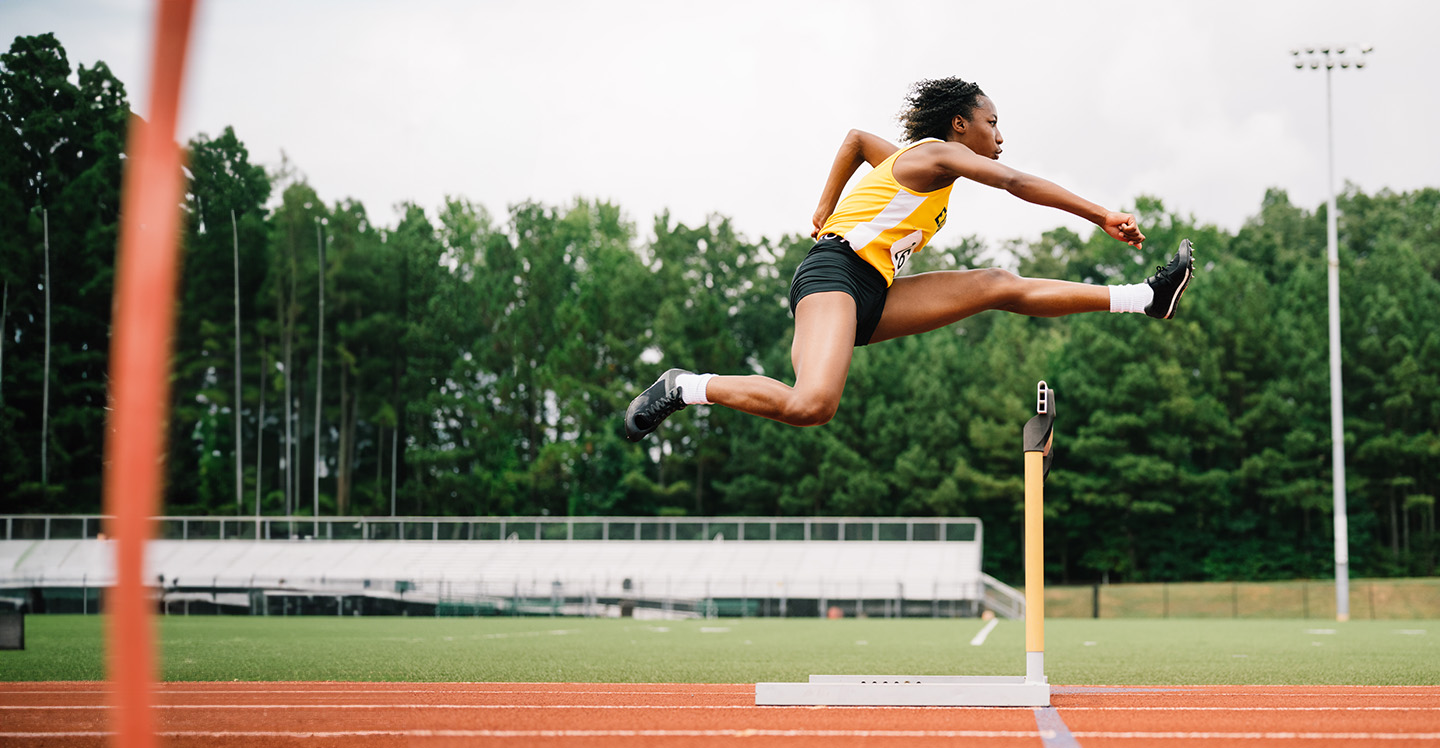 Female teen athlete jumps over hurdles on high school track.